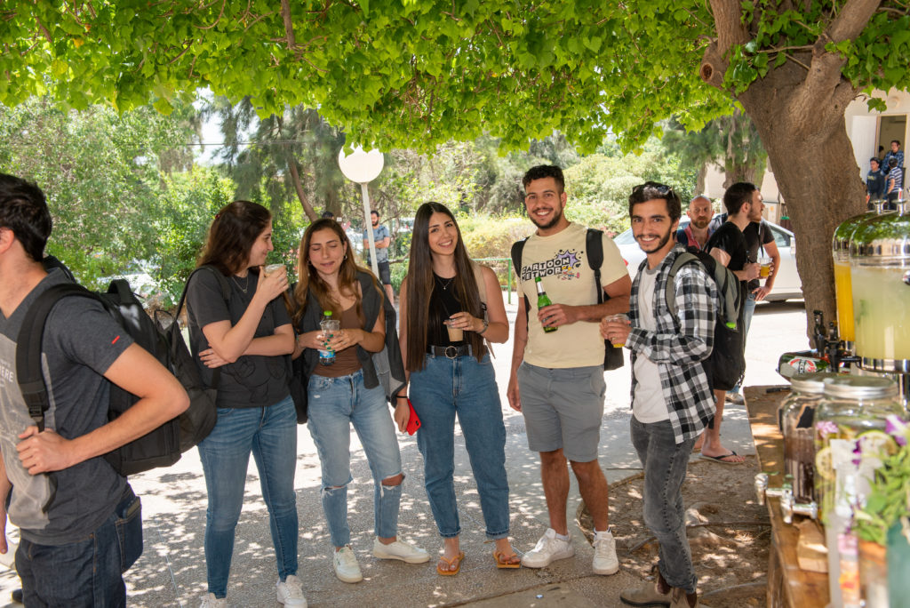 A group of 5 students, three women and two men, stand in a line and smile for the camera while socializing and standing under the trees outside.