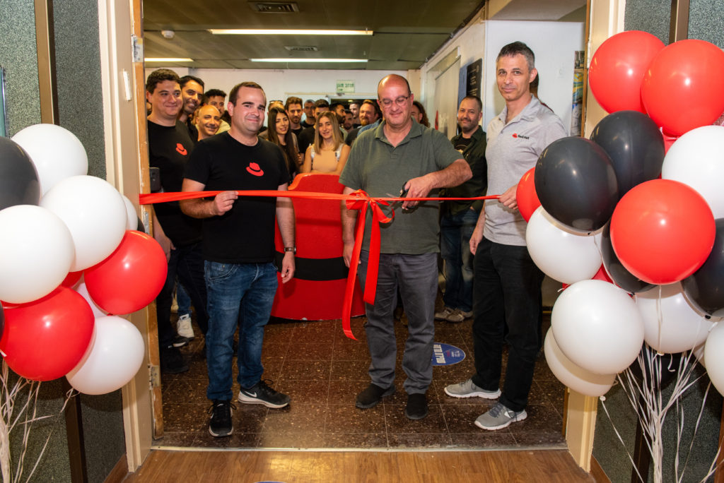 Red Hat associates and Tel-Hai Academic college staff and students watch as a man cuts a ceremonial red ribbon. There are red, white, and black ballons indicating the festive nature of the event.