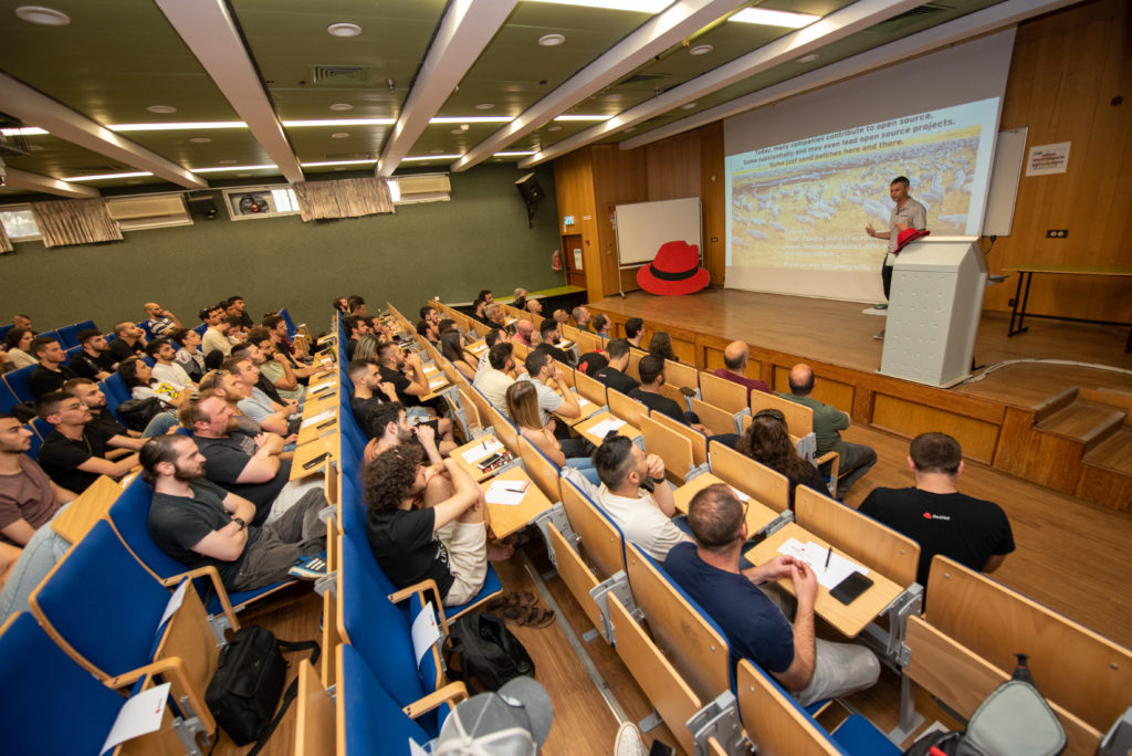 Long rows of students and guests seated in a lecture hall watch a man giving a presentation on open source culture and contributing to open source projects.